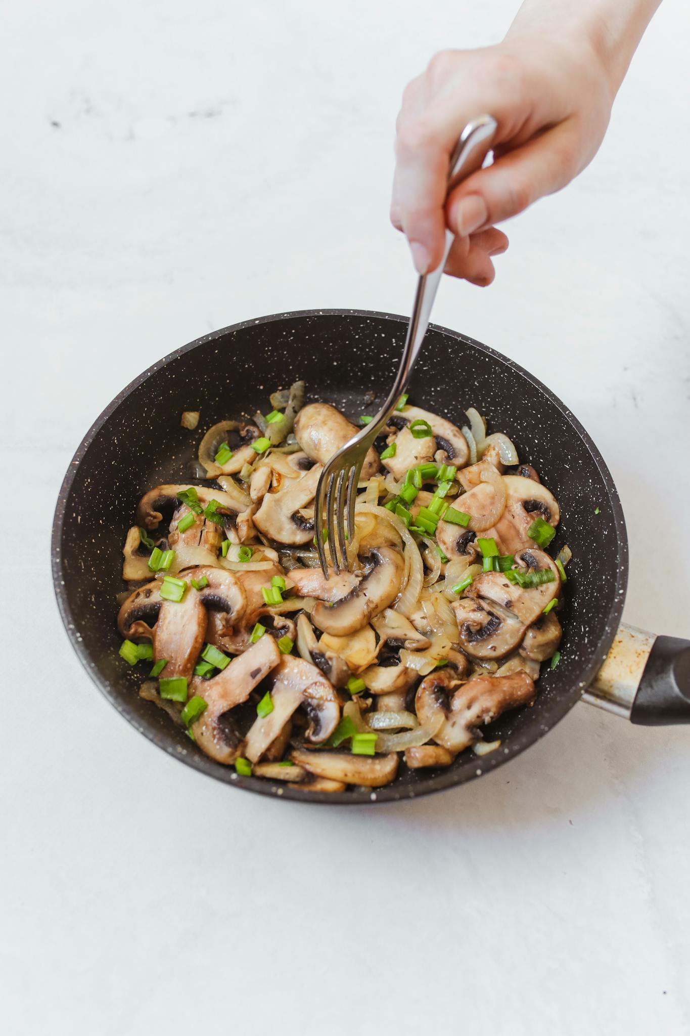 A frying pan full of sautéed mushrooms and vegetables with a hand stirring using a fork.