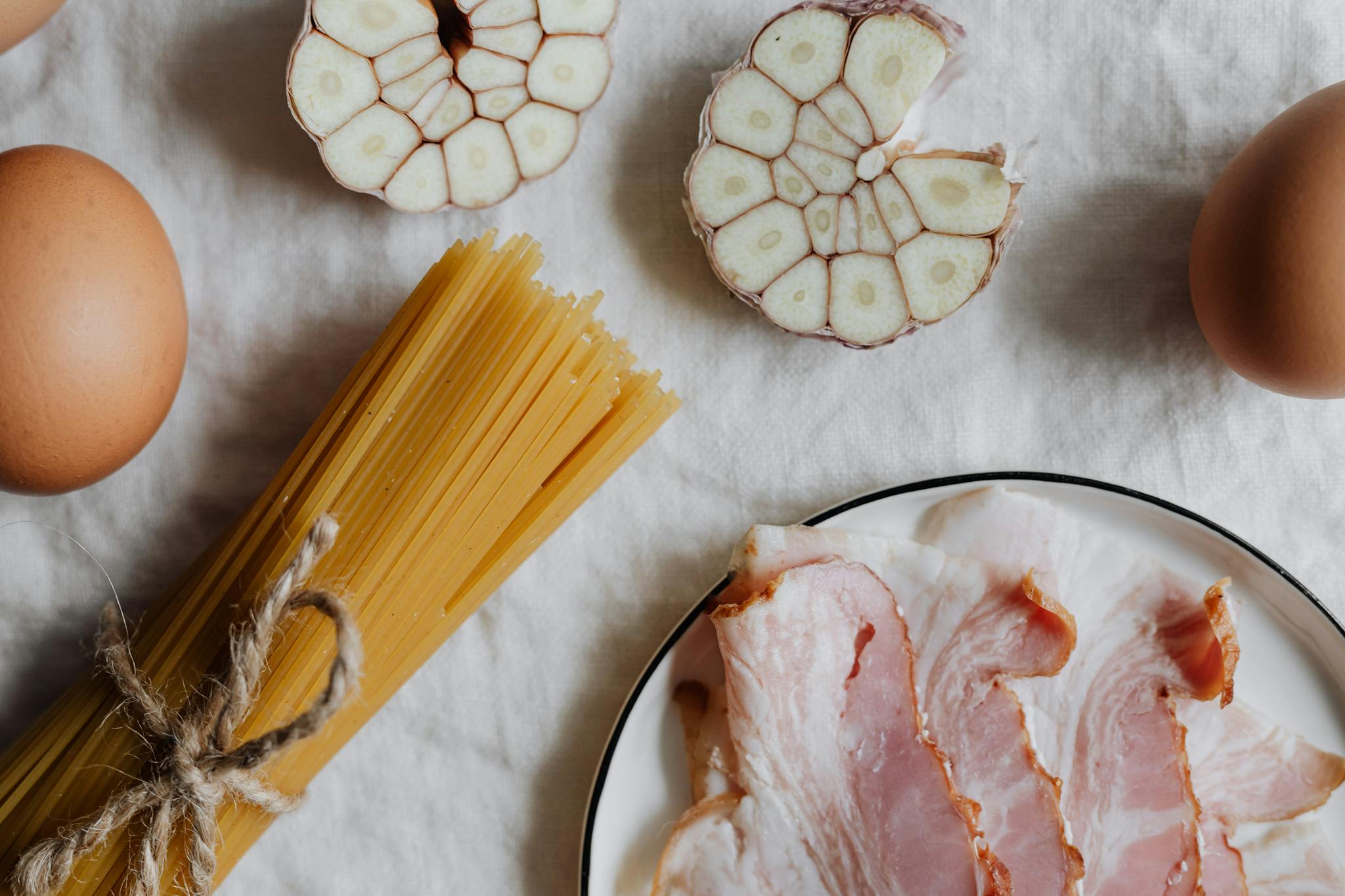 Flat lay of ingredients for classic carbonara: pasta, bacon, eggs, garlic on a white background.